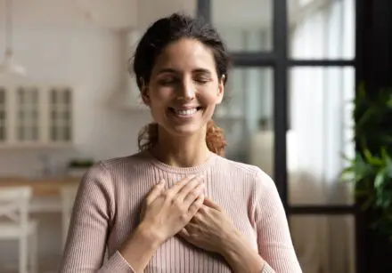 Dark haired woman with eyes closed, smiling, with her hands on her heart.