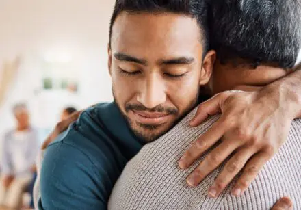 Father and son hugging, one facing us. Contented.