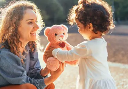 Curly haired woman giving a teddy bear to her little daughter outdoors in the park - family lifestyle concept.