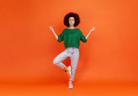 Portrait of woman with Afro hairstyle wearing green sweater with mudra gesture hands up, closed eyes, meditating standing in yoga position. Indoor studio shot isolated on orange background.
