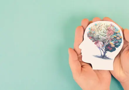 Hands holding paper head with human brain made of a tree with flowers. Set against a pastel aqua background.