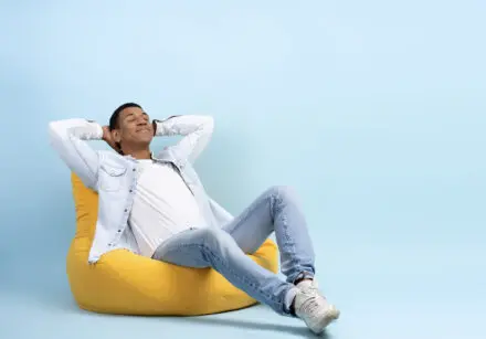 Relaxed cheerful man relaxing on a yellow bean bag chair with a light sky blue blue wall in the background. He's wearing light blue jeans and denim jacket and white t-shirt.