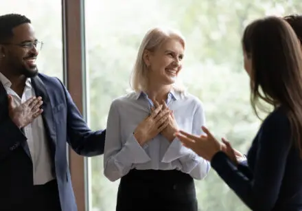 Middle-aged businesswoman receiving praises from multi ethnic colleagues. She is wearing a pale blue blouse and black skirt, has medium length blond hair, and is smiling broadly.