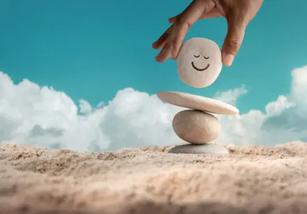 A hand putting a pebble with a smile drawn on it onto a small pile of three pebbles - flat on the bottom, a round one in the middle, and a flat one on top. The smile pebble is being placed on top of this one. It's on a beach and there's a blue sky in the background.