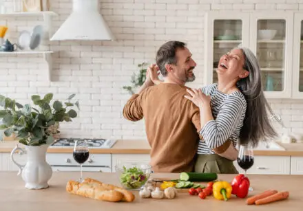 Happy cheerful middle-aged mature couple dancing together in the kitchen, preparing food. On the worktop are vegetables (red and yellow pepper), cucumber, tomatoes, mushrooms, carrots and salad. The salad is in a glass bowl. There's also two baguettes and two glasses of red wine.