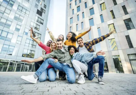 Group of friends bunched together, happy, each with one arm outstretched to their sides, with broad smiles on their faces. Kneeling on concrete paving stones with college campus buildings in the background.