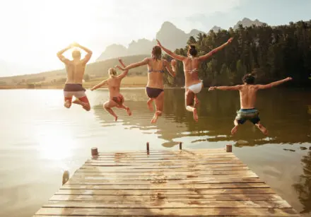 Portrait of young friends jumping from jetty into lake. It's a sunny day and they're all wearing swimming costumes.