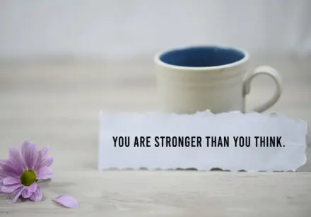 A cream coloured cup on a table with a pink daisy flower beside it. Resting on the cup is a torn off piece of paper with the words, 'You are stronger than you think' typed on it.