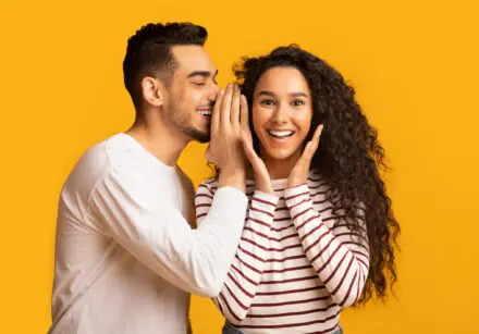 Young man sharing news with his excited girlfriend. He's whispering something in her ear. They both look happy. They're standing against a bright yellow background.