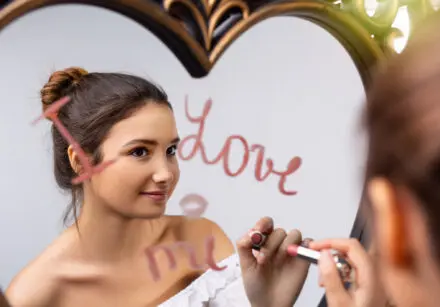 Portrait of a young girl with dark hair looking in a heart-shaped mirror and writing "I love me" onto it in pink lipstick.