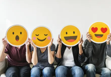 Four people sitting on a sofa, each holding a large human face-sized emoji that covers their face. From left to right the emojis are: surprise, contentment, happiness, love (that is, two red love hearts over the eyes). Each person is casually dressed in jeans and a t-shirt.
