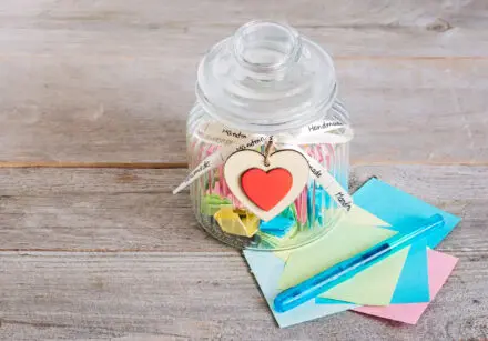 Glass jar with handmade wooden heart decoration and ribbon sitting on a wooden plank. There's folded pieces of pastel coloured paper inside. There's a pen beside the jar on top of unfolded pieces of the pastel coloured paper.