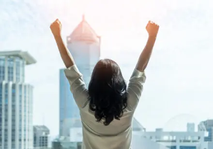 Woman with arms in the air in a depiction of success. She's looking out towards a city from a window high up.