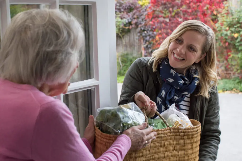 Female neighbor helping a senior woman with shopping. They are at the senior woman's door and the younger neighbour is handing her a basket of food.