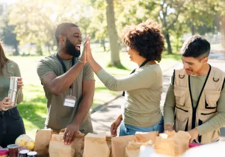 4 charity volunteers in a park, packing brown paper gift bags. Two in the middle are giving each other a high five. It's a bright, sunny day, and they're all smiling. There's green grass and trees in the background. They're all wearing earth-green coloured tops.