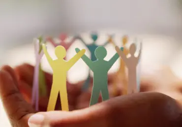 Mental Health Awareness Week 2025. Image of hands holding a cardboard cut-out of people in a circle holding hands. Each person is in a different colour of card.