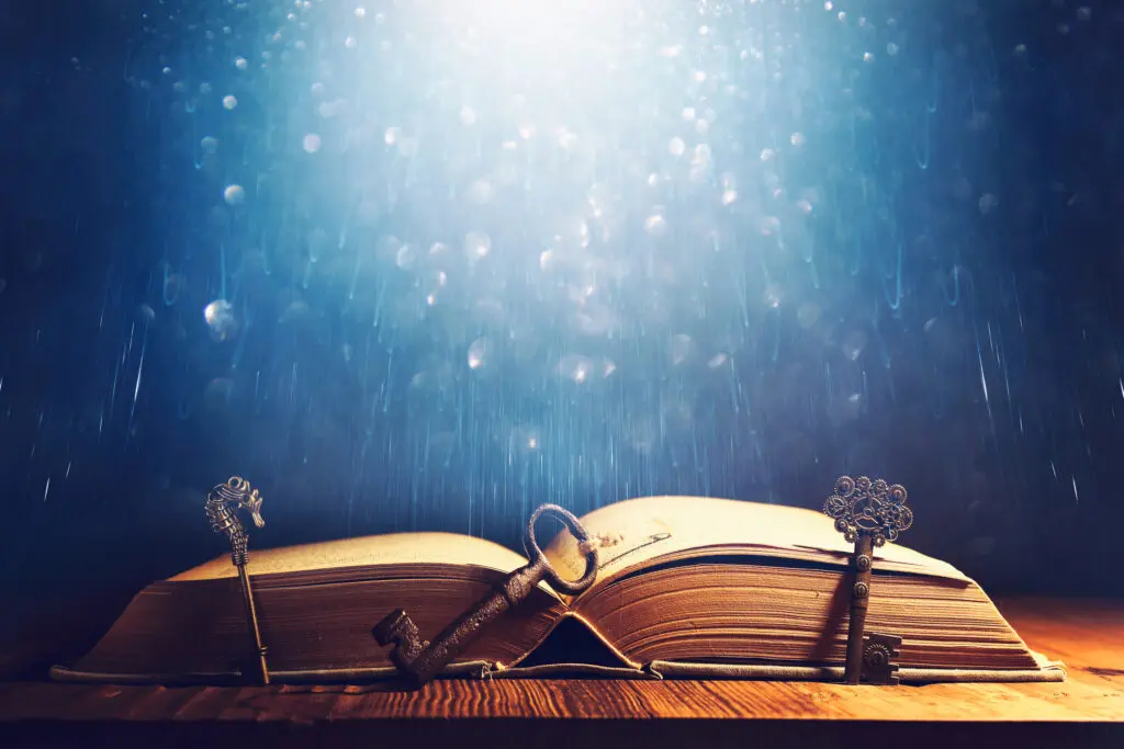 Magical looking image of an old, antique open book on a table with three old keys leaning against it. The background is blue with sparkles through it.