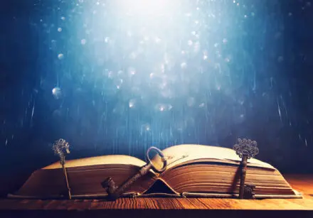 Magical looking image of an old, antique open book on a table with three old keys leaning against it. The background is blue with sparkles through it.
