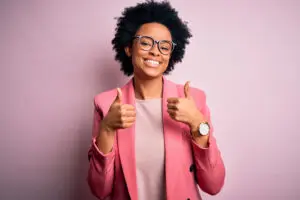 Young beautiful African American afro businesswoman with curly hair wearing pink jacket success sign doing positive gesture with hand, thumbs up smiling and happy. Cheerful expression and winner gesture. The wall behind her is baby-pink.