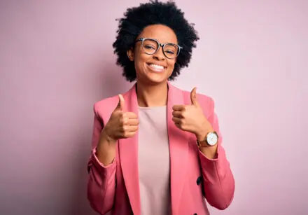 Young beautiful African American afro businesswoman with curly hair wearing pink jacket success sign doing positive gesture with hand, thumbs up smiling and happy. Cheerful expression and winner gesture. The wall behind her is baby-pink.