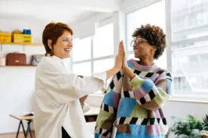 Tow women smiling and giving each other a High Five. The woman on the left is wearing a white blouse and the woman on the right is wearing a multi coloured jumper of coloured squares - blue, pink, olive green, while, and black. They're in a well-lit, spacious office.
