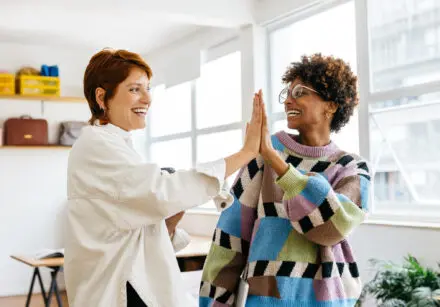 Tow women smiling and giving each other a High Five. The woman on the left is wearing a white blouse and the woman on the right is wearing a multi coloured jumper of coloured squares - blue, pink, olive green, while, and black. They're in a well-lit, spacious office.
