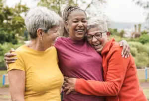Three woman around 50-60 years old hugging each other in a park. They're all laughing. The one on the left is wearing a mustard-colour t-shirt, the one in the middle is wearing a pink t-shirt, and the one on the right is wearing an orange sweatshirt.