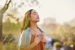 Young woman with hand on chest breathing in fresh air in a beautiful garden during sunset. She is Latino, has dark brown long hair, is wearing a brown vest with a great open blouse on top.
