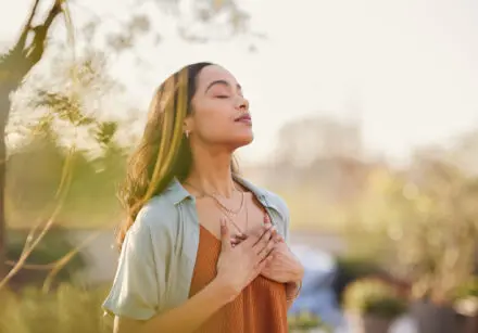Young woman with hand on chest breathing in fresh air in a beautiful garden during sunset. She is Latino, has dark brown long hair, is wearing a brown vest with a great open blouse on top.