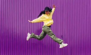 Young woman with dark hair and black cap, wearing a yellow top, khaki green cargo pants and white sneakers, against a purple background. She's a hip hop dancer and is in the air with arms outstretched and leaping.