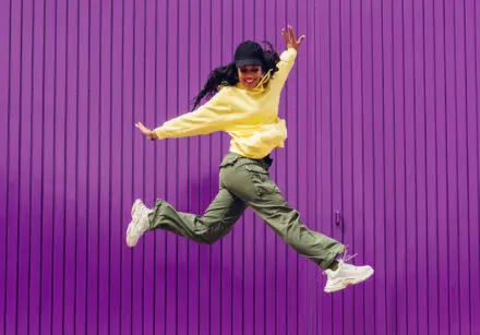 Young woman with dark hair and black cap, wearing a yellow top, khaki green cargo pants and white sneakers, against a purple background. She's a hip hop dancer and is in the air with arms outstretched and leaping.