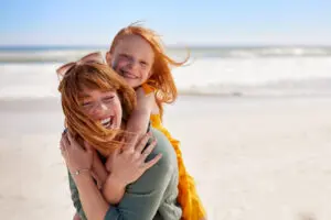 Laughing mother with daughter piggyback on her back, having fun at the beach. The sea is in background.