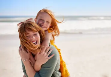 Laughing mother with daughter piggyback on her back, having fun at the beach. The sea is in background.