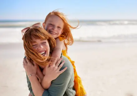 Laughing mother with daughter piggyback on her back, having fun at the beach. The sea is in background.
