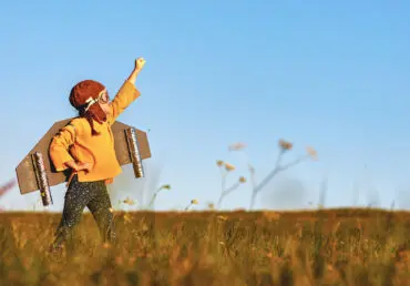 Child playing with wooden wings strapped to his back. He's wearing an aviator mask and has one fist pointed into the air. He is wearing dark trousers with stars all over them and a mustard top. He is in a field and the sky is pure light blue.