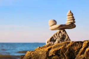 Stack of zen stones in balance at seashore. There's a large flat one on the bottom that represents the long part of a see saw, with smaller piles of zen stones at either end. The 'see saw' is balanced on the tip of a triangular stone. It's on a rock with sea and blue sky in the background.