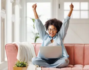 Young smiling African American woman smiling with arms in the air, rejoicing in a victory. She's sitting cross-legged on a salmon-coloured sofa and has a laptop on her lap. The room behind is blurred out, but is brightly lit. We can see a tall green plant and a back door with a 6 pane window at the top.