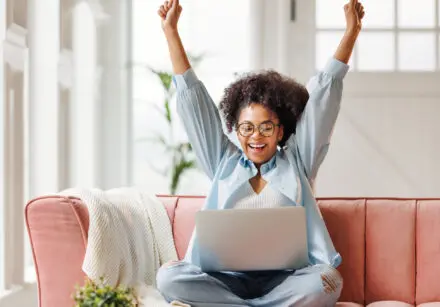 Young smiling African American woman smiling with arms in the air, rejoicing in a victory. She's sitting cross-legged on a salmon-coloured sofa and has a laptop on her lap. The room behind is blurred out, but is brightly lit. We can see a tall green plant and a back door with a 6 pane window at the top.