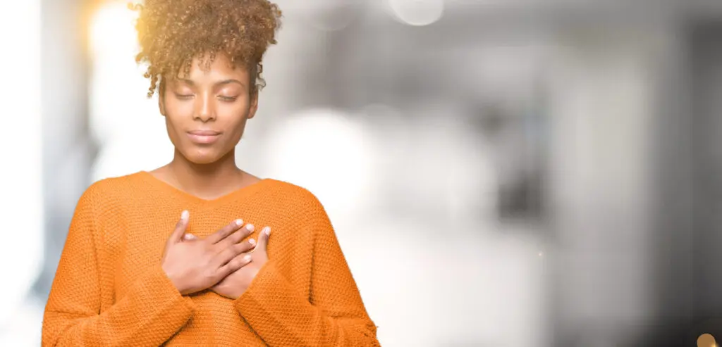 Young african american woman, wearing an orange jumper, over blurred background smiling with hands on her chest with closed eyes and grateful gesture on face.