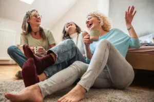 3 women around 40 years old laughing hysterically. They're sitting on the floor at the foot of a bed on a rug. They're all wearing jeans, have no shoes on, and are holding cups of tea or coffee. They're wearing light coloured tops - one is blue, one is white, and one is light green.