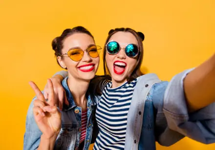 Two girls, happy, hugging facing the camera, wearing bright clothing and sunglasses. They're both wearing open blue denim jackets and striped tops. One has dark blue and white stripes and the other has read and white strips - both horizontal stripes. The girls on the left is smiling broadly and the girl on the right has a wide open mouth smile and is making a fun two finger peace gesture. They seem very happy and fun. The background is single tone mustard-yellow.