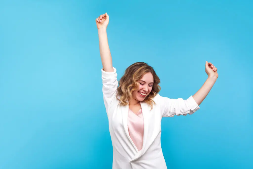 Young woman with light brown wavy hair looking joyful. She is wearing a white suit jacket with a pink top and the background is cyan. She is dancing - she has one arm straight up and the other is half way up, as if in the middle of a dance move. She is very happy.