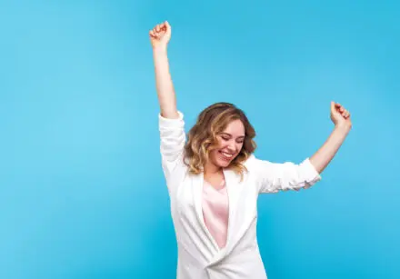 Young woman with light brown wavy hair looking joyful. She is wearing a white suit jacket with a pink top and the background is cyan. She is dancing - she has one arm straight up and the other is half way up, as if in the middle of a dance move. She is very happy.
