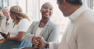 A man and woman in a well lit room, by the window, shaking hands as if meeting for the first time. The picture shows others meeting in the background so may be a meet-and-greet event or something similar. The woman is black and is wearing a grey-blue open jacket with white blouse. She is smiling. The man is wearing a cream jumper with white shirt collar showing, and is looking towards the woman.