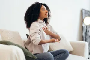 Young woman practicing breathing. She is sitting in a meditation position and has one hand on her heart and the other just in front of her tummy, with the palm up. She has dark shoulder length hair and is wearing a cream hooded top and blue jeans. She is on a white sofa with a white well behind her. The room is brightly lit.