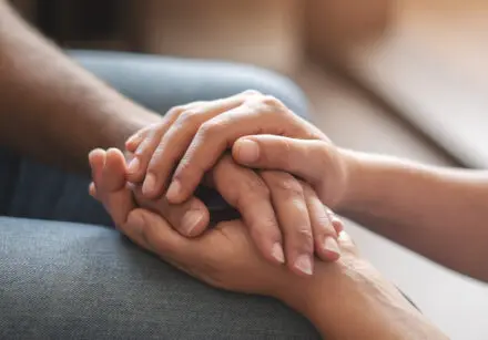 Closeup shot of two people holding hands in comfort. One is comforting the other. The hands are on the lap of the person being comforted and, from the angle, it appears that the comforter is on their knees.