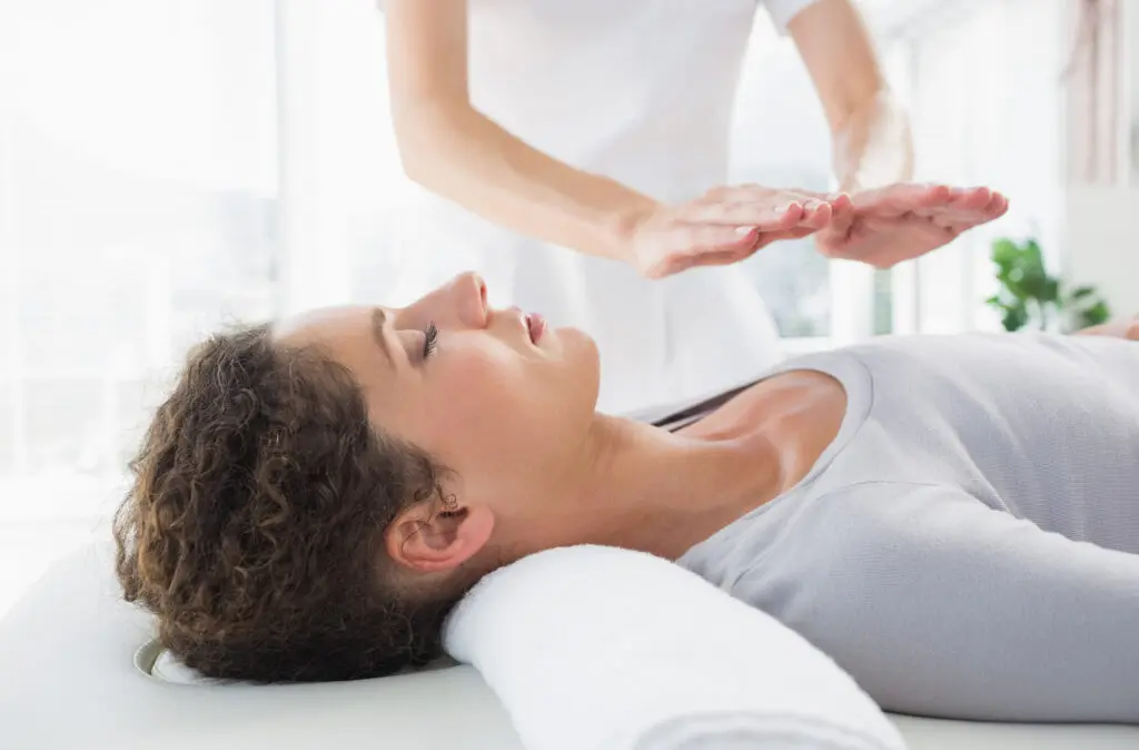 Woman on a therapy bed receiving reiki. A female practitioner stands with her hands over the chest area of the client. The rom is well lit with a large window in the background. The practitioner is wearing a white uniform with short sleeves. The client has a great top and has short dark hair.