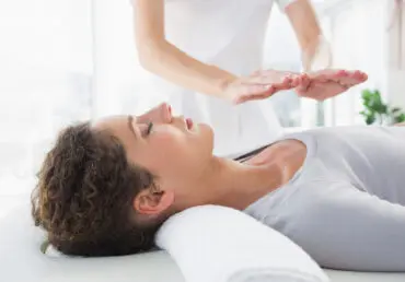 Woman on a therapy bed receiving reiki. A female practitioner stands with her hands over the chest area of the client. The rom is well lit with a large window in the background. The practitioner is wearing a white uniform with short sleeves. The client has a great top and has short dark hair.