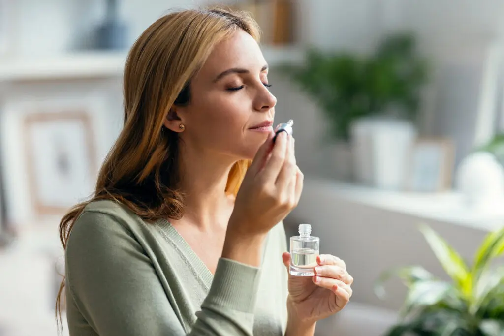 Woman with light brown hair with her eyes closed taking a sniff of an essential oil. She has the bottle in her left hand and holds the dropper to her nose with her right hand. She is wearing an olive green top. The background is blurred out, but it's a well lit room with what looks like plants in the background.
