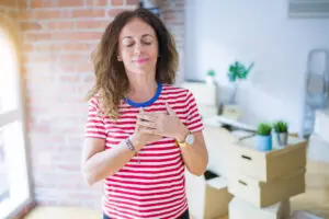 A woman in her 40s with her eyes closed, breathing, with her hands over her heart. She has wavy, shoulder-length brown hair, and is wearing a red and white horizontally striped t-shirt. She is standing in a well-lit room. An office with a desk and boxes is blurred out in the background, as well as an arched door with a red brick wall.